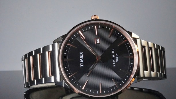 A sophisticated wristwatch with a black dial and rose gold markers, featuring the brand name 'Timex' and 'Sapphire Crystal' on the face. The stainless steel strap complements the minimalist and elegant design, with a small date window displaying the number two.