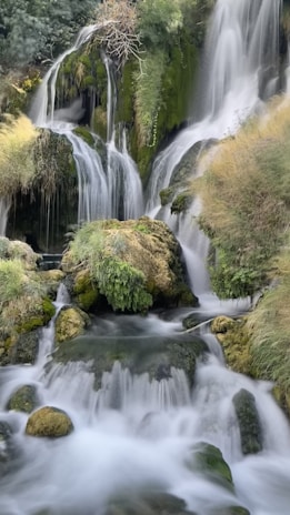 A serene waterfall cascading over moss-covered rocks, surrounded by lush greenery and foliage. The water flows smoothly, creating a gentle, calming effect.