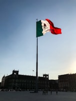 A large flagpole with the Mexican flag waves prominently against a clear sky. The horizon features historic buildings with colonial architecture, showing intricate details and silhouettes against the setting sun.