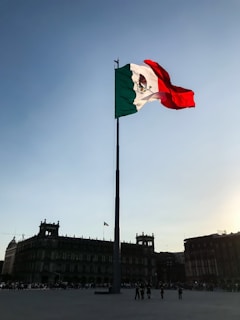 A large flagpole with the Mexican flag waves prominently against a clear sky. The horizon features historic buildings with colonial architecture, showing intricate details and silhouettes against the setting sun.