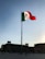 A large flagpole with the Mexican flag waves prominently against a clear sky. The horizon features historic buildings with colonial architecture, showing intricate details and silhouettes against the setting sun.