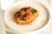 A plate of food featuring a crispy base topped with mashed orange spread and garnished with roasted cherry tomatoes, fresh dill, and other vegetables. The dish is presented on a simple, light-colored ceramic plate, situated on a white marble surface.