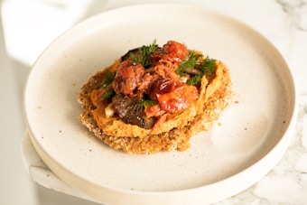 A plate of food featuring a crispy base topped with mashed orange spread and garnished with roasted cherry tomatoes, fresh dill, and other vegetables. The dish is presented on a simple, light-colored ceramic plate, situated on a white marble surface.
