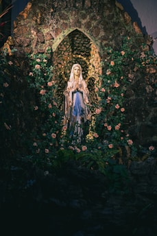 A religious statue of a woman in a blue and white robe is surrounded by a grotto of stone and green foliage. The statue is encircled by a crown of golden roses, and natural light falls softly, highlighting the delicate features and serene expression.