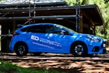 A blue sports car is parked outdoors in front of a structure under construction, surrounded by scaffolding and trees. The car features branding and logos related to driving school services. Sunlight filters through the leaves, casting shadows on the car and the ground.