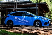 A blue sports car is parked outdoors in front of a structure under construction, surrounded by scaffolding and trees. The car features branding and logos related to driving school services. Sunlight filters through the leaves, casting shadows on the car and the ground.