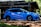 A blue sports car is parked outdoors in front of a structure under construction, surrounded by scaffolding and trees. The car features branding and logos related to driving school services. Sunlight filters through the leaves, casting shadows on the car and the ground.