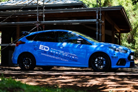 A blue sports car is parked outdoors in front of a structure under construction, surrounded by scaffolding and trees. The car features branding and logos related to driving school services. Sunlight filters through the leaves, casting shadows on the car and the ground.