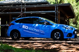 A blue sports car is parked outdoors in front of a structure under construction, surrounded by scaffolding and trees. The car features branding and logos related to driving school services. Sunlight filters through the leaves, casting shadows on the car and the ground.