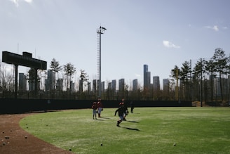 A baseball field with a few people walking on the green grass. The field is surrounded by a black fence on which a scoreboard is mounted. In the background, tall modern buildings and a few trees are visible under a clear blue sky.