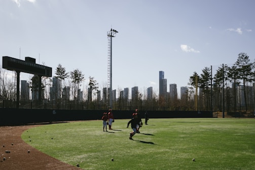 A baseball field with a few people walking on the green grass. The field is surrounded by a black fence on which a scoreboard is mounted. In the background, tall modern buildings and a few trees are visible under a clear blue sky.