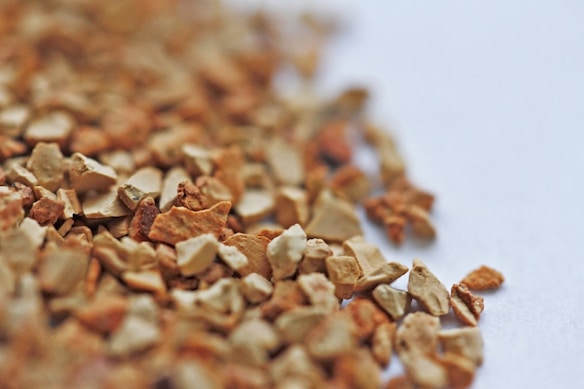 A close-up of small, irregularly shaped granules with a rough texture, predominantly in shades of brown and tan. They are scattered across a light surface, with a shallow depth of field that results in a blurred background.