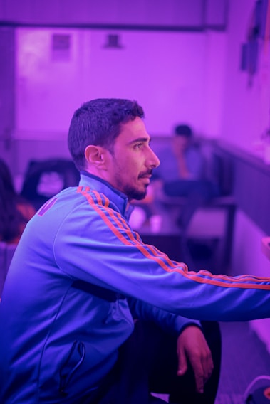 A man wearing a blue tracksuit with orange stripes is sitting in a room with a purple hue. The lighting gives the scene a relaxed and somewhat surreal atmosphere. In the background, another person is visible, partially blurred, seated on a sofa.