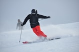 A skier in bright red pants is skillfully navigating down a snowy slope, leaving a trail of powdery snow behind. The skier is dressed in a black jacket and helmet, with protective goggles. In the background, another skier can be seen, also equipped with winter sports gear.