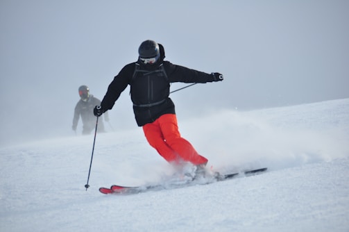 A skier in bright red pants is skillfully navigating down a snowy slope, leaving a trail of powdery snow behind. The skier is dressed in a black jacket and helmet, with protective goggles. In the background, another skier can be seen, also equipped with winter sports gear.
