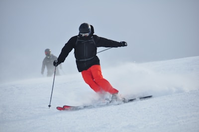 A skier in bright red pants is skillfully navigating down a snowy slope, leaving a trail of powdery snow behind. The skier is dressed in a black jacket and helmet, with protective goggles. In the background, another skier can be seen, also equipped with winter sports gear.