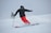 A skier in bright red pants is skillfully navigating down a snowy slope, leaving a trail of powdery snow behind. The skier is dressed in a black jacket and helmet, with protective goggles. In the background, another skier can be seen, also equipped with winter sports gear.