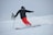 A skier in bright red pants is skillfully navigating down a snowy slope, leaving a trail of powdery snow behind. The skier is dressed in a black jacket and helmet, with protective goggles. In the background, another skier can be seen, also equipped with winter sports gear.