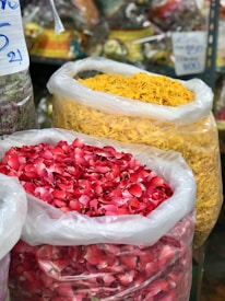 Large plastic bags filled with vibrant red and yellow flower petals are prominently displayed, suggesting a market setting with other colorful items in the blurred background.