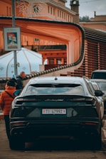 A black luxury car is parked on a street with an elaborate architectural structure in the background. People are walking on the sidewalk, dressed in winter clothing. The scene conveys an urban setting with an artistic and modern design element in the architecture.