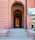 A long, arched brick corridor with multiple layers of arches extending into the distance, creating an effect of depth and perspective. The corridor is flanked by large brick walls with a signboard on the right side, advertising tribal textiles, tribal paintings, metal crafts, and e-commerce. There are steps leading up to the corridor, and part of a building's facade with windows is visible on the right.