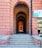 A long, arched brick corridor with multiple layers of arches extending into the distance, creating an effect of depth and perspective. The corridor is flanked by large brick walls with a signboard on the right side, advertising tribal textiles, tribal paintings, metal crafts, and e-commerce. There are steps leading up to the corridor, and part of a building's facade with windows is visible on the right.