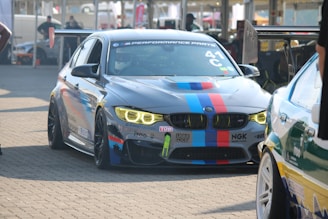 A sleek BMW race car in a pit area with multi-colored racing stripes and sponsor decals. The car features prominent halo headlights and a large rear wing, surrounded by other vehicles and racing equipment.