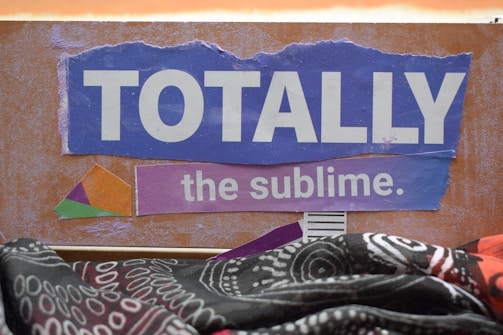 A textured artwork features the words 'TOTALLY the sublime.' on a purple torn paper against a brown background. There is a geometric colorful shape to the left, and a patterned fabric with black, white, and red designs draped across the bottom.