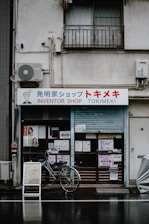 A storefront labeled 'Inventor Shop Tokimeki' with various signs and advertisements placed around the entrance. A bicycle is parked in front of the shop. The building shows signs of aging, with some electrical equipment and an air conditioning unit attached to the wall.