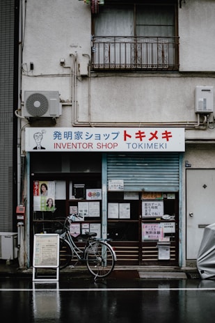 A storefront labeled 'Inventor Shop Tokimeki' with various signs and advertisements placed around the entrance. A bicycle is parked in front of the shop. The building shows signs of aging, with some electrical equipment and an air conditioning unit attached to the wall.