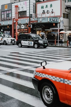 A city street scene in Japan with several cars, including a black taxi and an orange taxi, moving across a wet, rainy crosswalk. Pedestrians, some holding umbrellas, can be seen in the background in front of various shops and buildings, including ones with Japanese signage.