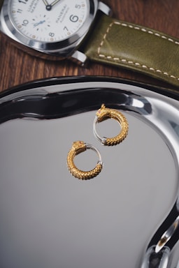 A pair of intricately designed gold hoop earrings with a textured pattern resembling scales is placed on a shiny metallic surface. Nearby, a luxury wristwatch with a green leather strap and a white dial rests on a wooden surface, exhibiting a classic and elegant style.