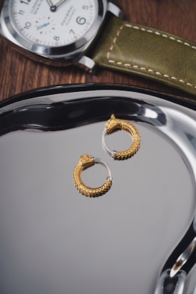A pair of intricately designed gold hoop earrings with a textured pattern resembling scales is placed on a shiny metallic surface. Nearby, a luxury wristwatch with a green leather strap and a white dial rests on a wooden surface, exhibiting a classic and elegant style.
