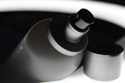 A close-up view of a spray bottle with its cap removed. The bottle has a sleek, matte black finish, and the nozzle is prominently displayed. The background contrasts with bright highlights, giving a modern and polished appearance.
