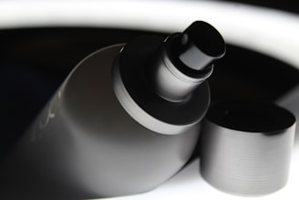 A close-up view of a spray bottle with its cap removed. The bottle has a sleek, matte black finish, and the nozzle is prominently displayed. The background contrasts with bright highlights, giving a modern and polished appearance.