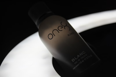 A silhouette of a perfume bottle with the brand name 'one' and the word 'BLANC' on it, set against a dark background with a soft, glowing light source beneath it, creating a moody and elegant atmosphere.