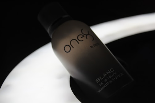 A silhouette of a perfume bottle with the brand name 'one' and the word 'BLANC' on it, set against a dark background with a soft, glowing light source beneath it, creating a moody and elegant atmosphere.