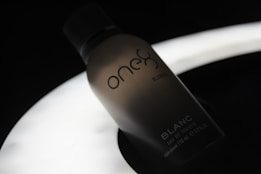 A silhouette of a perfume bottle with the brand name 'one' and the word 'BLANC' on it, set against a dark background with a soft, glowing light source beneath it, creating a moody and elegant atmosphere.