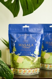 A package of 'Masala Blend' Indian Masala Chai tea on a wooden surface, surrounded by lush green leaves. The packaging is predominantly blue with intricate designs and text in gold and white, featuring an artistic landscape illustration at the bottom.