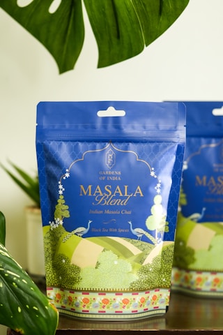A package of 'Masala Blend' Indian Masala Chai tea on a wooden surface, surrounded by lush green leaves. The packaging is predominantly blue with intricate designs and text in gold and white, featuring an artistic landscape illustration at the bottom.