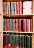 A wooden bookshelf filled with three tiers of vintage-style books, predominantly featuring hardcovers with ornate, embossed gold designs. The books are mostly shades of red, green, and black, with some blue and brown spines. One book on the second shelf to the right is noticeably different with a white cover that has colorful abstract designs and readable text promoting health.