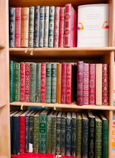 A wooden bookshelf filled with three tiers of vintage-style books, predominantly featuring hardcovers with ornate, embossed gold designs. The books are mostly shades of red, green, and black, with some blue and brown spines. One book on the second shelf to the right is noticeably different with a white cover that has colorful abstract designs and readable text promoting health.