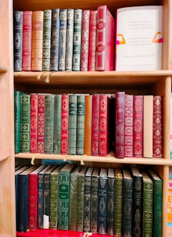 A wooden bookshelf filled with three tiers of vintage-style books, predominantly featuring hardcovers with ornate, embossed gold designs. The books are mostly shades of red, green, and black, with some blue and brown spines. One book on the second shelf to the right is noticeably different with a white cover that has colorful abstract designs and readable text promoting health.