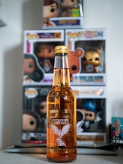 A bottle labeled 'Potion du Phoenix' is placed in front of several boxed figurines. The bottle is amber-colored with a label featuring a white phoenix. The figurines appear to be collector's items displayed in their original packaging.