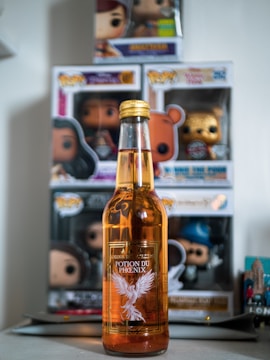 A bottle labeled 'Potion du Phoenix' is placed in front of several boxed figurines. The bottle is amber-colored with a label featuring a white phoenix. The figurines appear to be collector's items displayed in their original packaging.