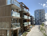 Modern apartment buildings with wooden facades and glass balconies, set along a waterfront promenade. The architecture features clean lines and open designs, offering an airy and spacious vibe. Plants and small trees line the walkway, adding a touch of nature to the urban setting.