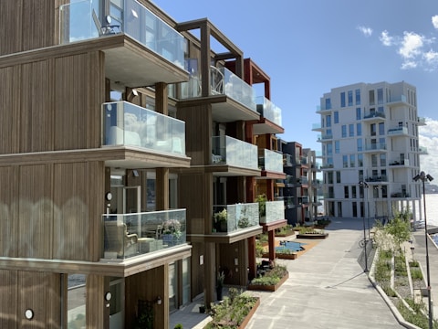 Modern apartment buildings with wooden facades and glass balconies, set along a waterfront promenade. The architecture features clean lines and open designs, offering an airy and spacious vibe. Plants and small trees line the walkway, adding a touch of nature to the urban setting.