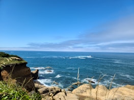 An ocean view with waves breaking against the shoreline rocks. The coastline includes rocky cliffs with green vegetation on the left, and the vast expanse of the blue sea under a clear sky with a few clouds.