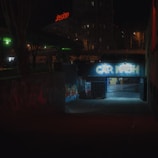 A dimly lit urban setting featuring a tunnel entrance with graffiti text 'CAR WASH' illuminated by harsh artificial lighting. Surrounding the entrance are dark structures with vibrant graffiti. In the background, there's a neon red sign atop a building and silhouettes of leafless trees against a night sky.