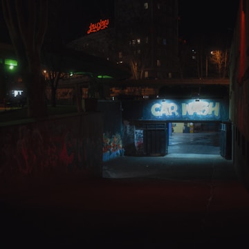 A dimly lit urban setting featuring a tunnel entrance with graffiti text 'CAR WASH' illuminated by harsh artificial lighting. Surrounding the entrance are dark structures with vibrant graffiti. In the background, there's a neon red sign atop a building and silhouettes of leafless trees against a night sky.